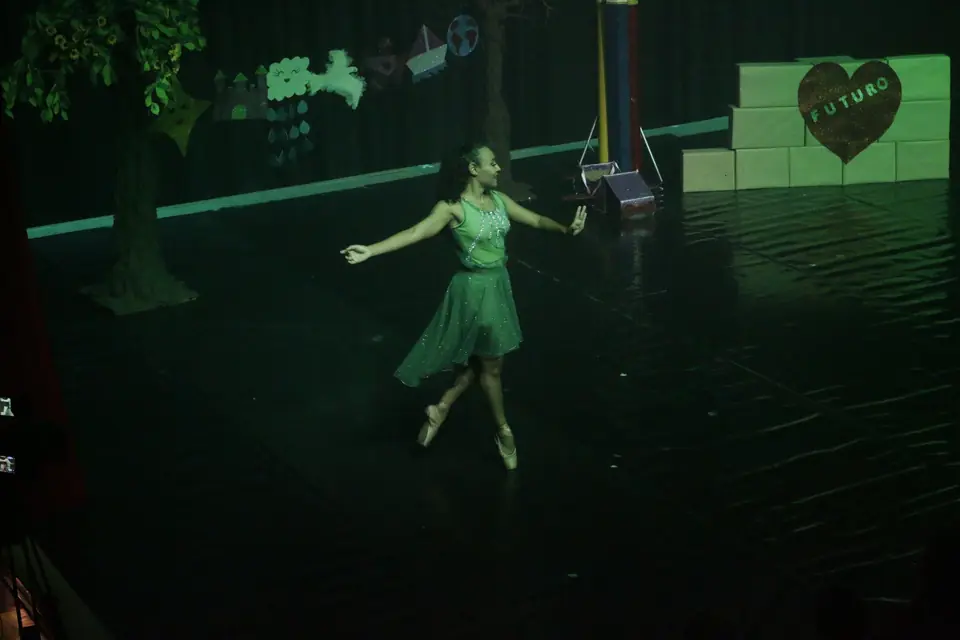 Bailarina de verde dança sozinha em palco amplo, com árvore e varal de desenhos no cenário.