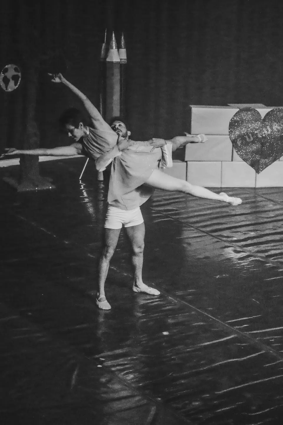Fotografia em preto e branco de um duo: bailarino sustenta bailarina em grande extensão de pernas.