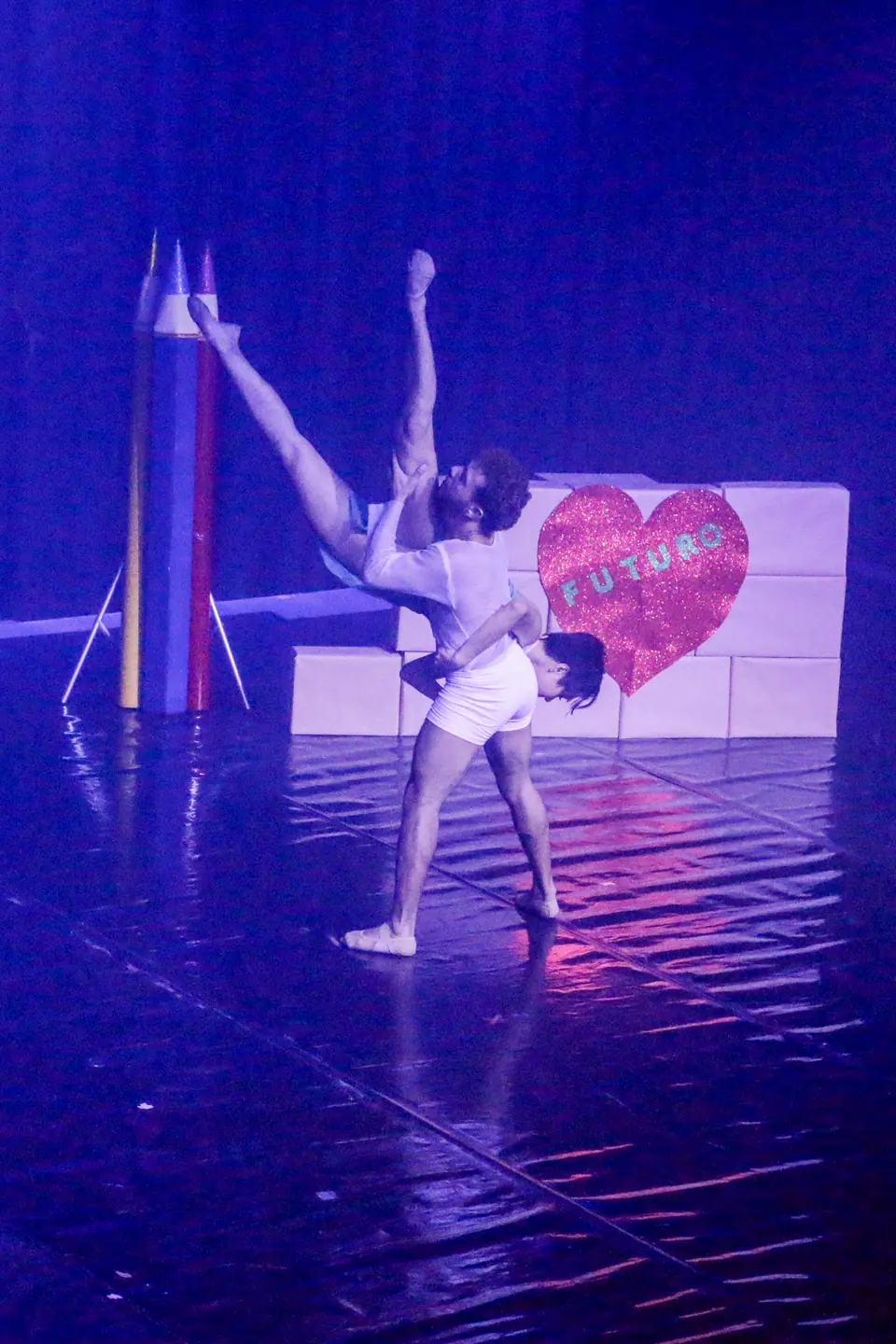 Bailarino ergue uma bailarina em arco diante de um muro de blocos com um coração onde se lê FUTURO, sob luz azul.