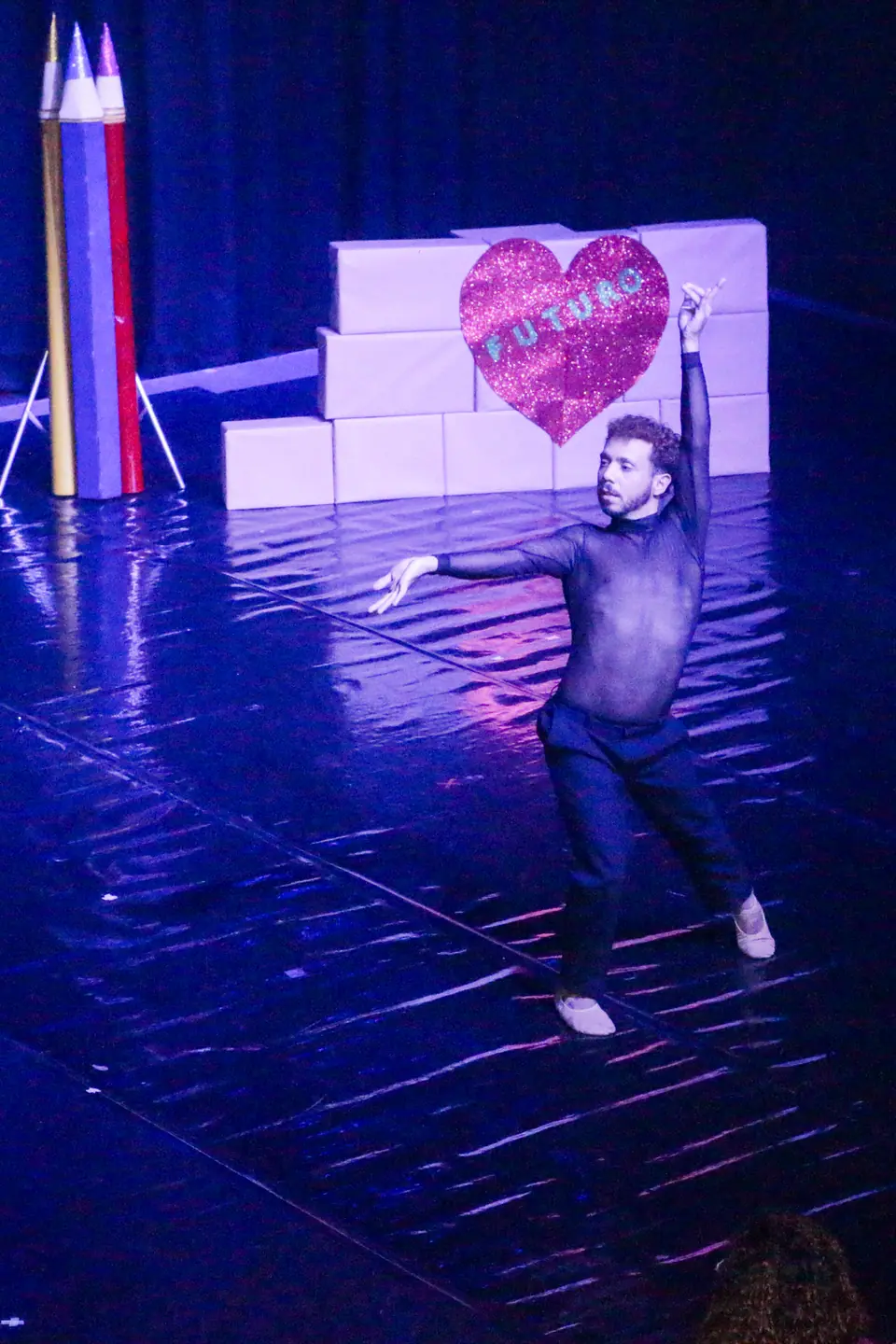Bailarino em solo com um braço erguido diante de lápis gigantes coloridos e do coração FUTURO.