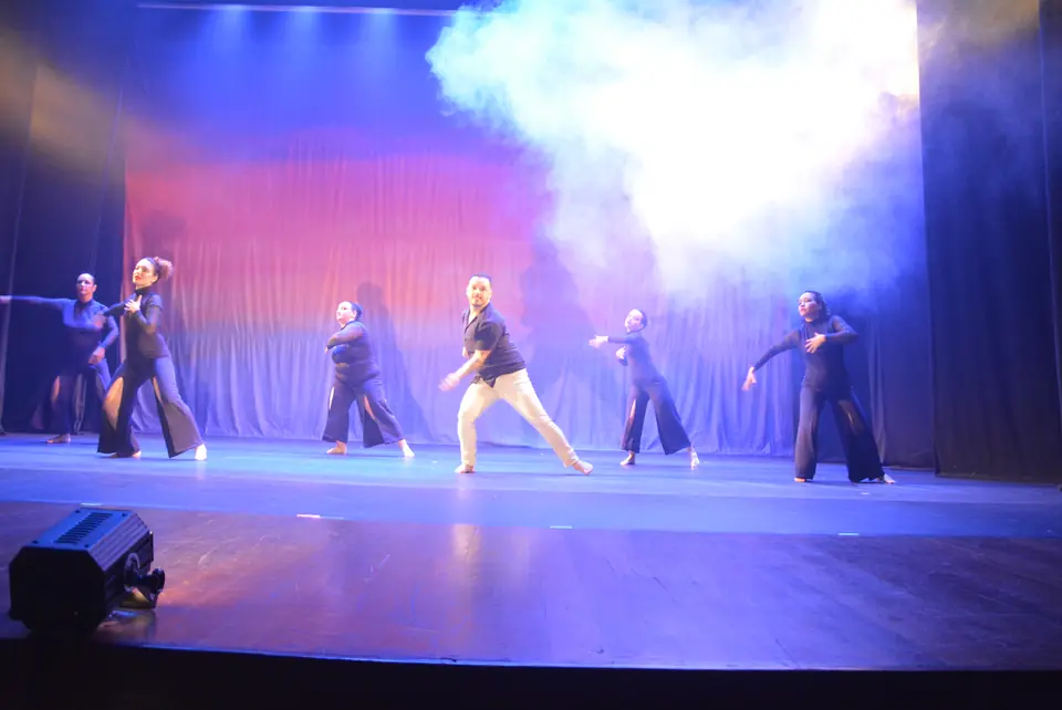 Grupo de bailarinos com figurinos escuros e claros espalhados pelo palco, com névoa e luz azul.