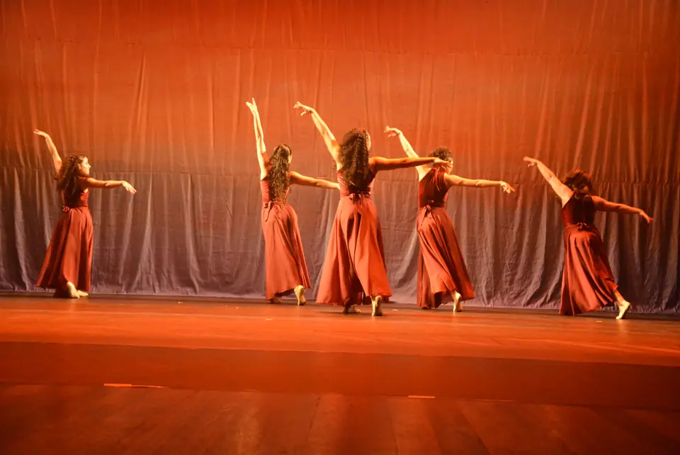Bailarinas com vestidos vermelhos em linha diagonal, braços erguidos, sob luz laranja intensa.
